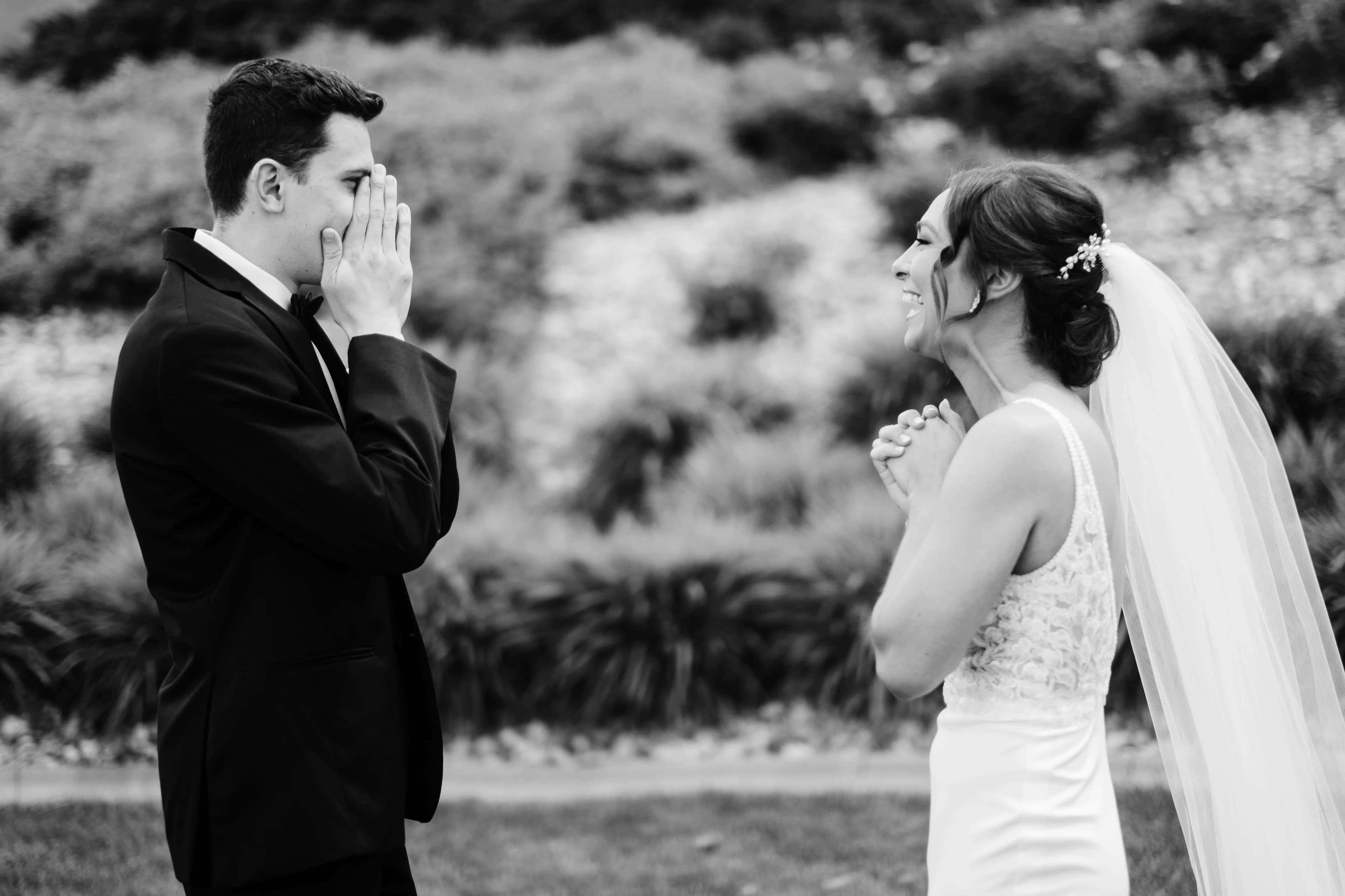 A groom covers his face with both hands, overwhelmed during a first look, while the bride faces him laughing with her hands clasped, photographed outdoors before the ceremony.