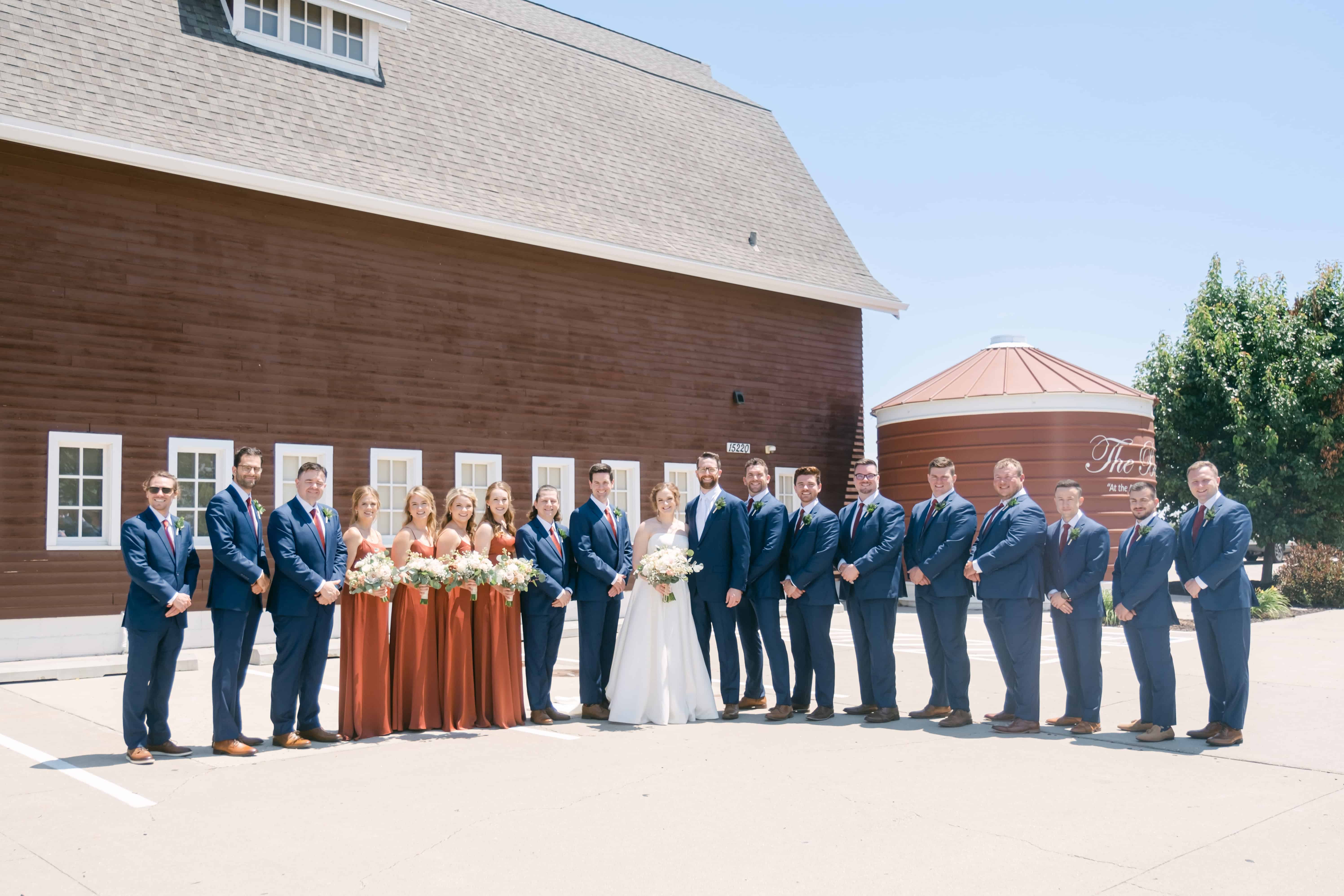 The entire wedding party lined up in one frame outside a red barn venue, the bride and groom at the center, bridesmaids in rust dresses holding blush bouquets, and a long row of groomsmen in navy suits on either side.