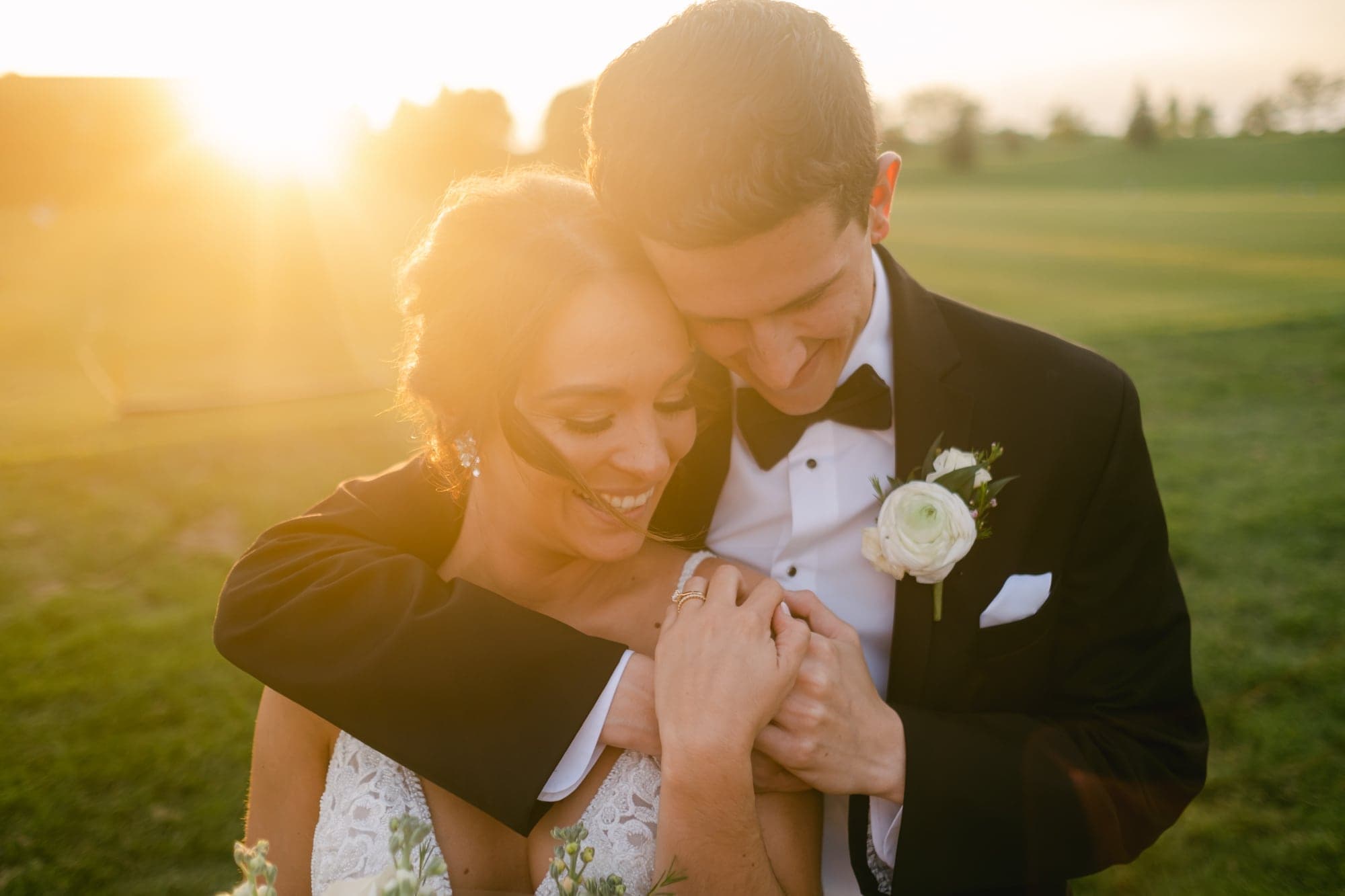 A groom wraps his arms around a bride from behind in an open field at golden hour, the setting sun flaring low on the horizon behind them. Taken around 8:20 PM in late May in Omaha, Nebraska, 22 minutes before sunset.