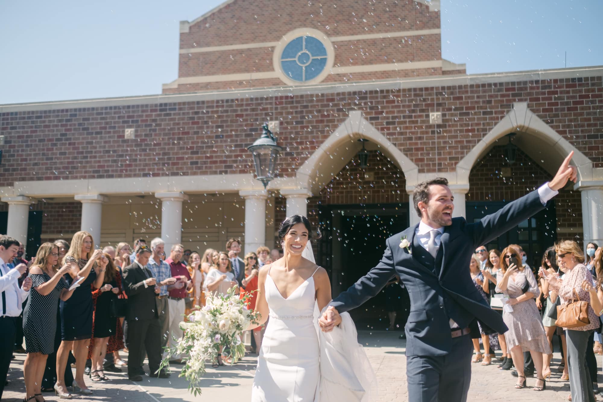 A bride and groom exit a brick church into a bubble send-off, smiling as guests line both sides blowing bubbles in the sunshine.
