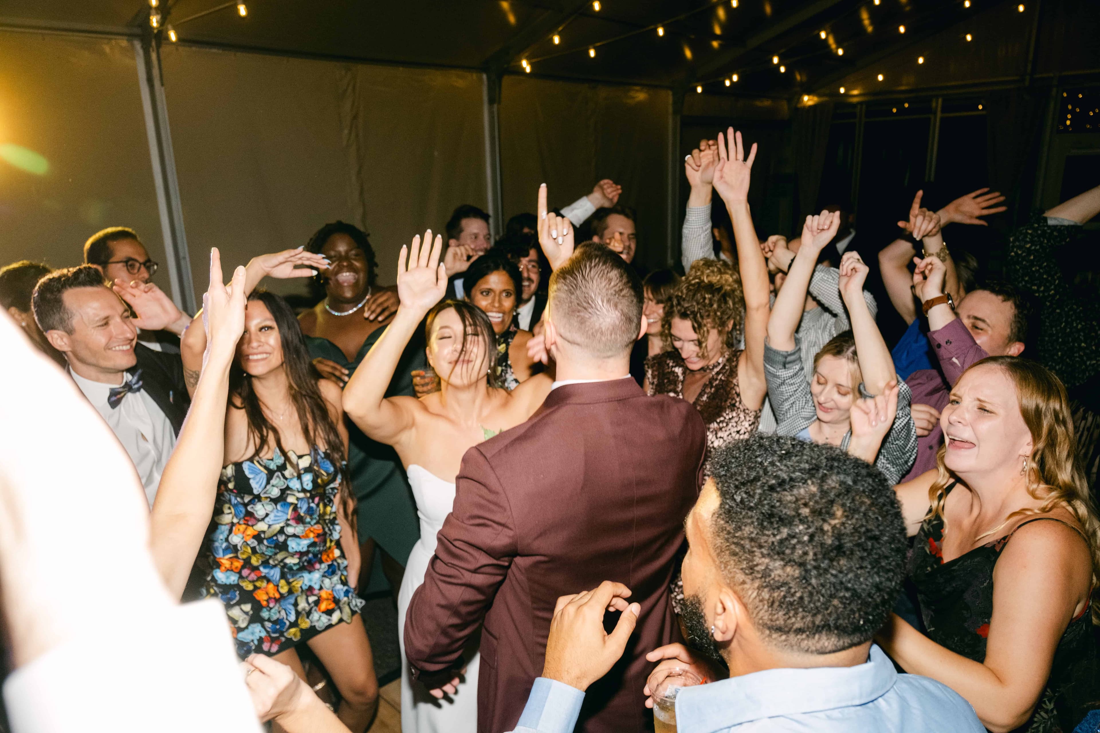 A packed dance floor under a tent at night, the bride in a white dress laughing with her arms raised beside the groom in a burgundy suit, seen from behind at the center as guests crowd in around them with hands in the air under string lights.