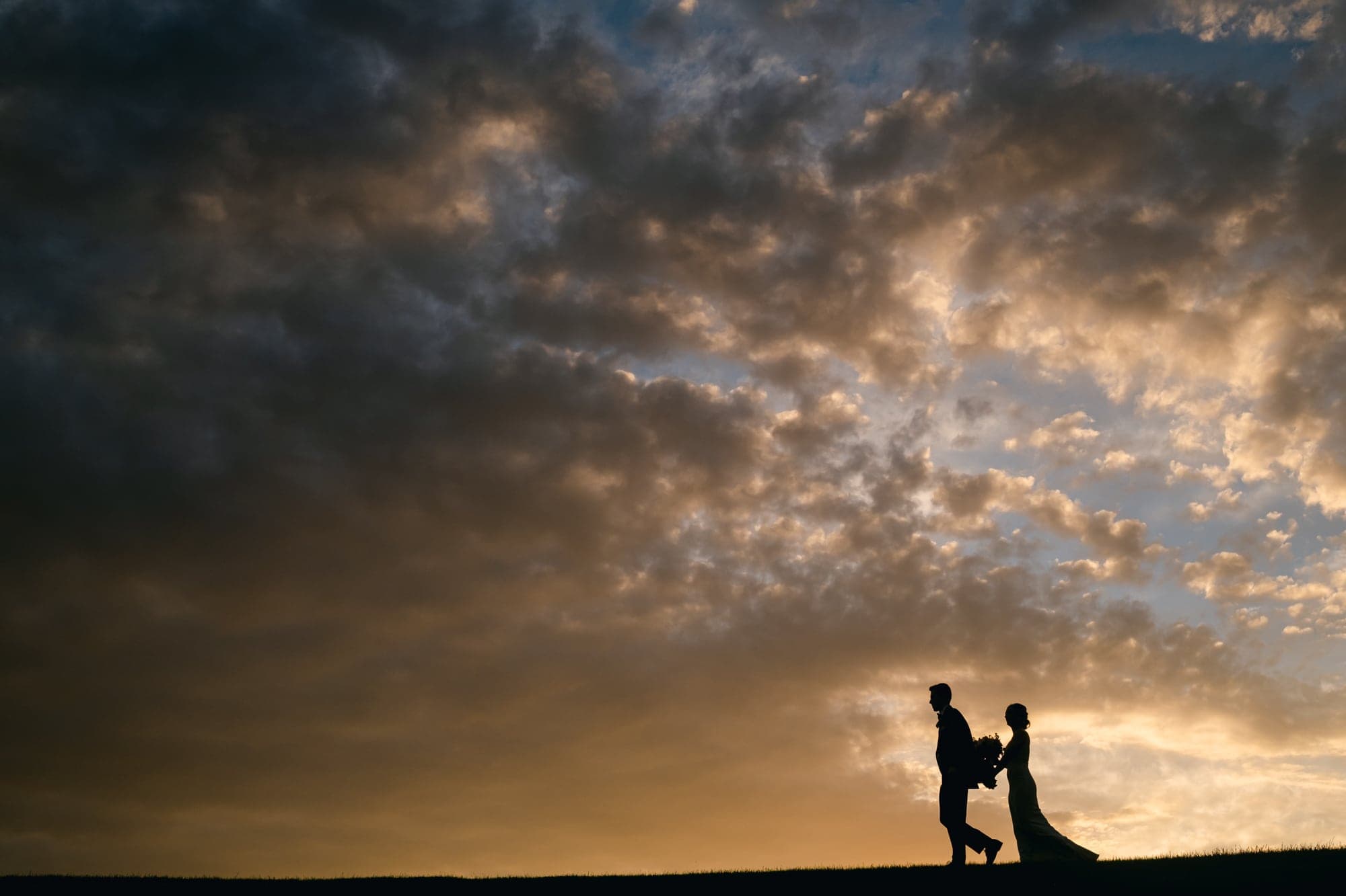 A bride and groom walk along a hilltop, silhouetted against a dramatic golden-hour sky at sunset.