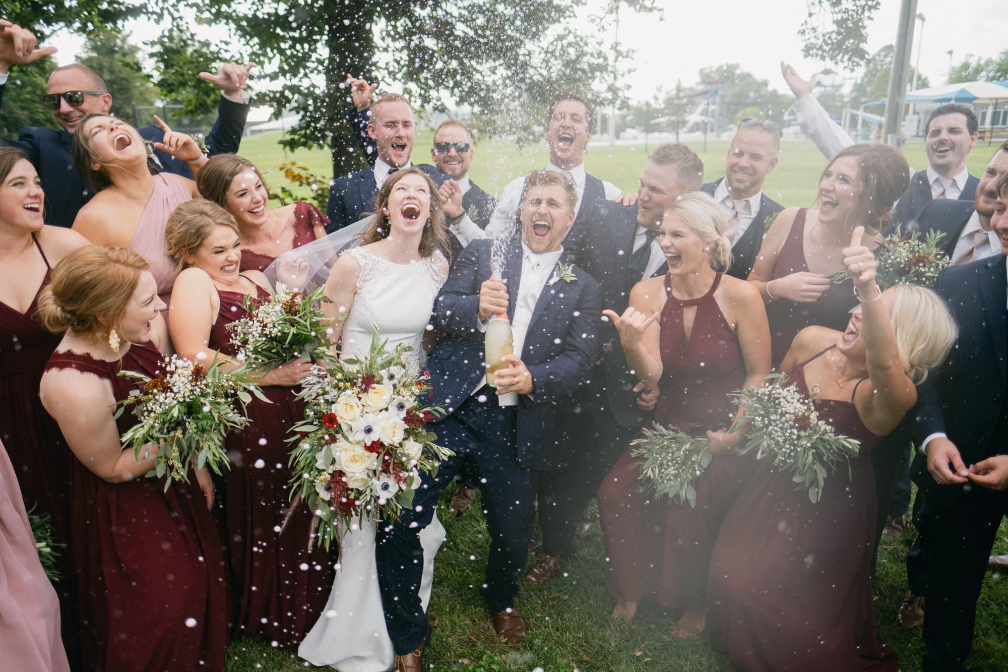 The full wedding party crowded together under a tree as the groom pops a bottle of champagne, spray flying, the bride laughing beside him with bridesmaids in burgundy and groomsmen in navy all around.