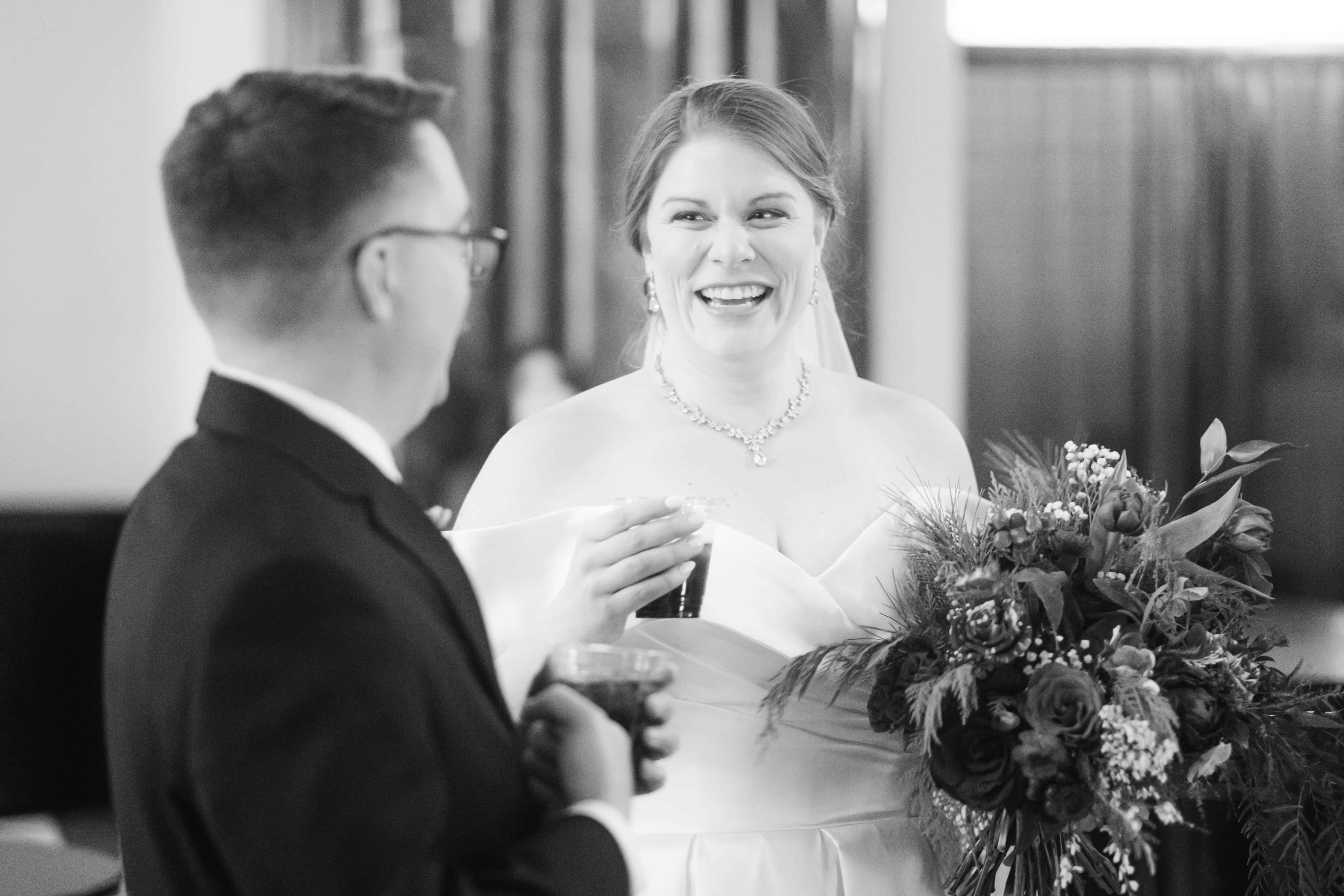 A bride laughs mid-conversation at cocktail hour, drink in one hand and bouquet still in the other, facing the groom as he stands with her, both holding glasses.