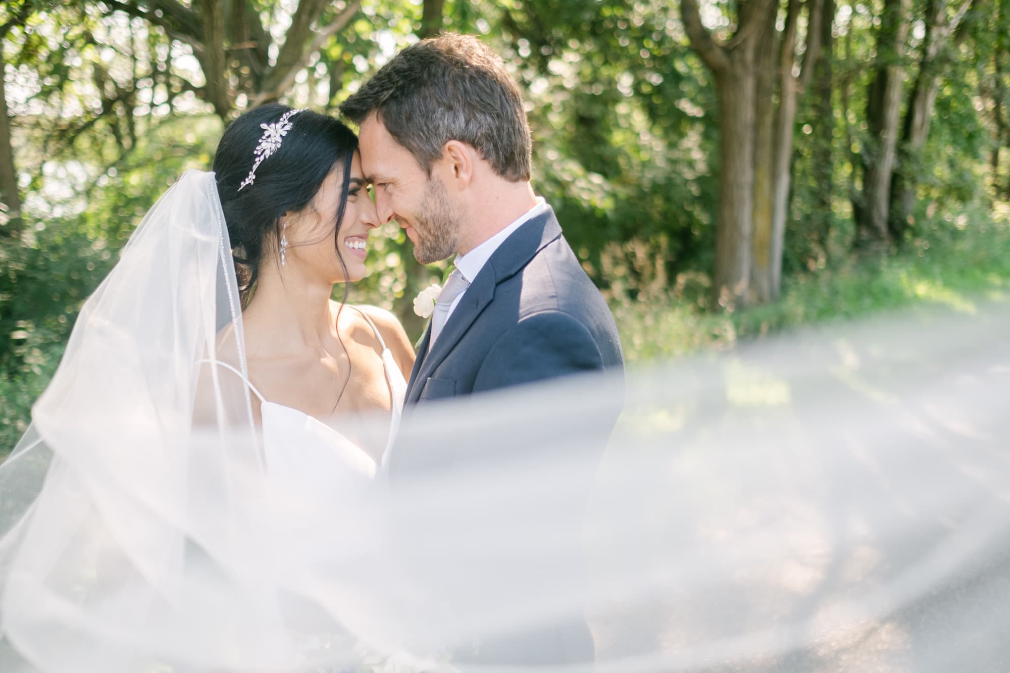 A bride and groom touch foreheads and smile during a portrait among sunlit trees, the bride's veil sweeping across the foreground.