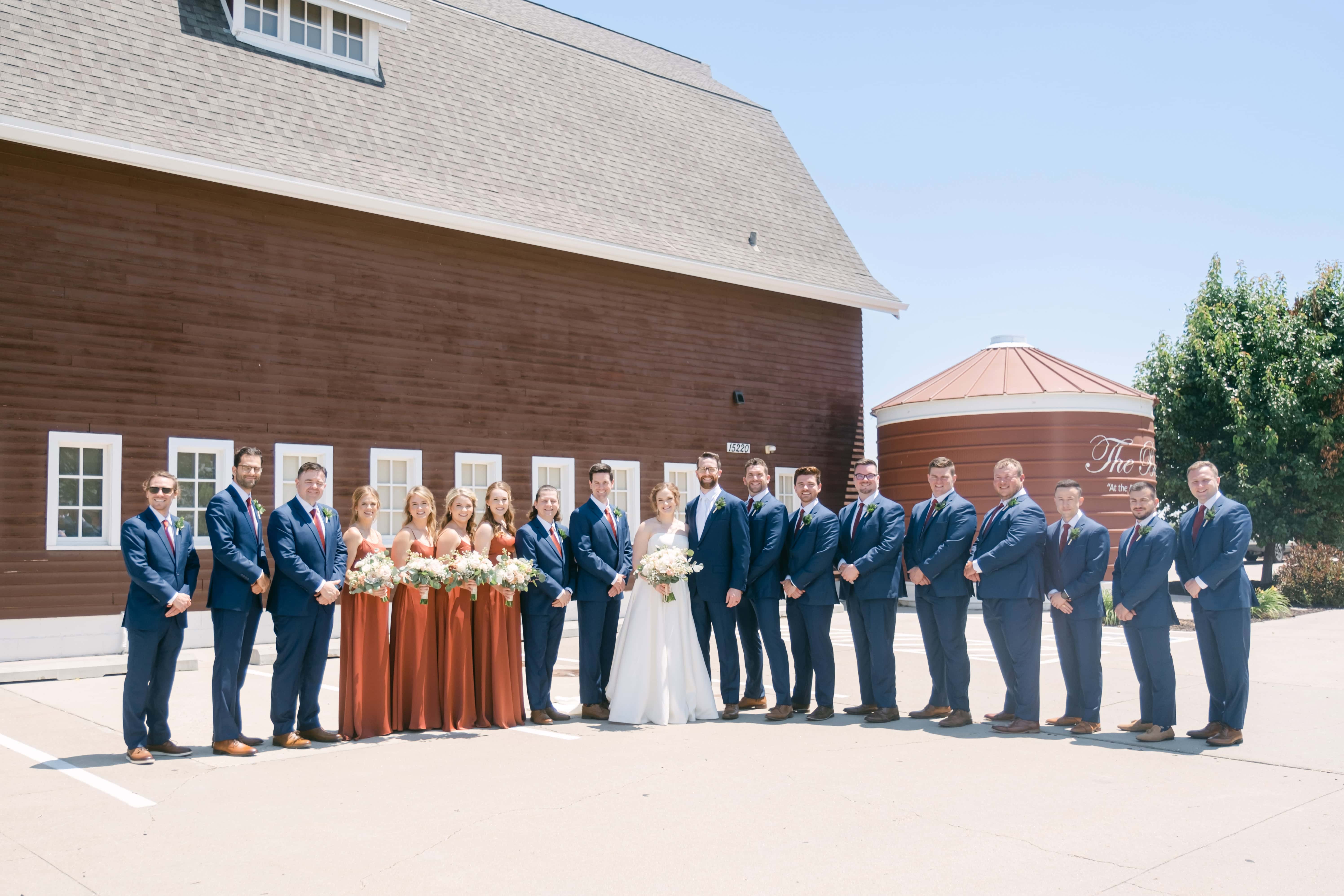 A large wedding party lined up shoulder to shoulder outside a red barn venue, the bride and groom at the center, groomsmen in navy suits and bridesmaids in rust dresses.